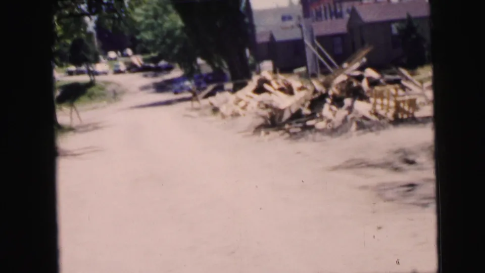 Beachside path transitioning to a residential street in what is described as Camden, NJ, circa 1964; trees, a low-rise building, and a lumber or debris pile visible.