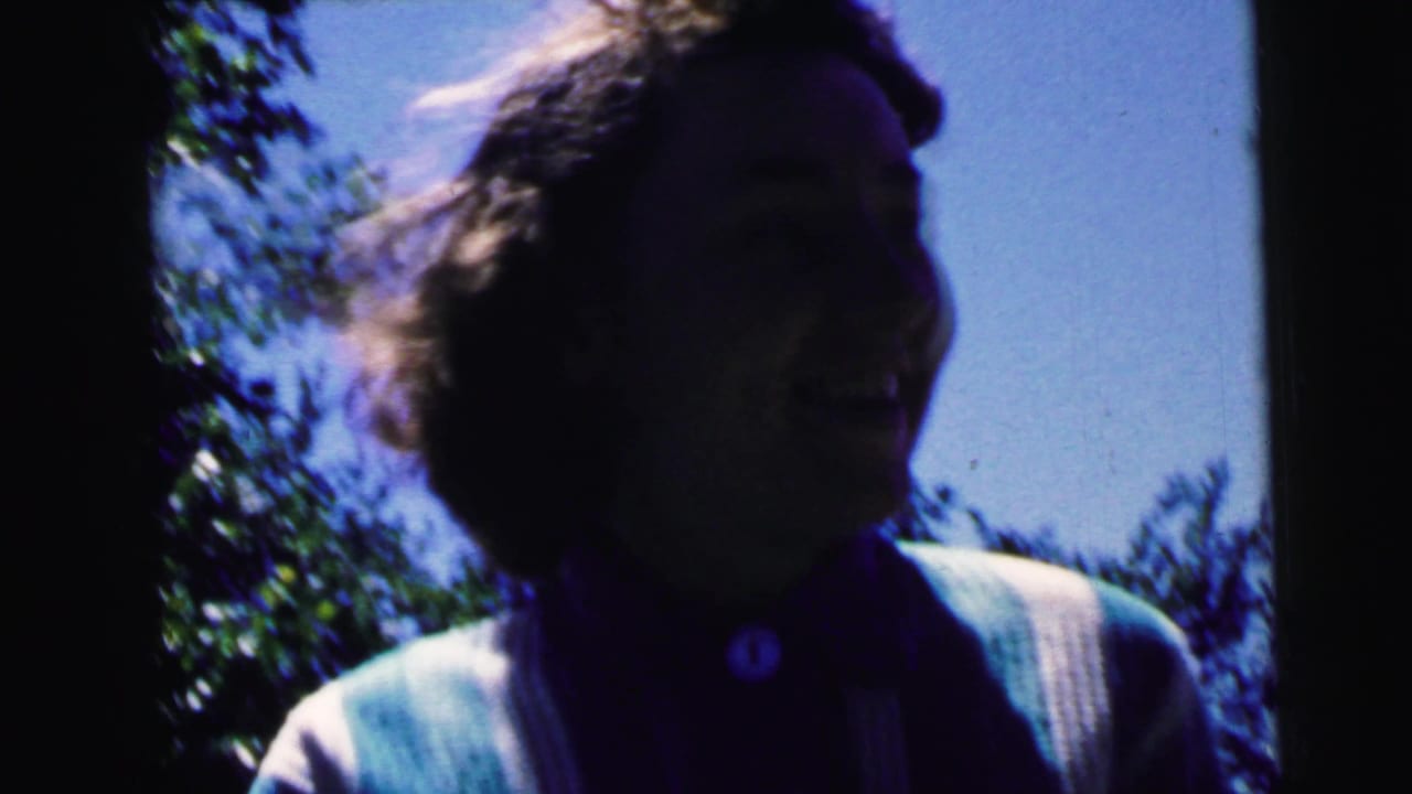 Laughing woman with curly hair and light cardigan, framed against backyard sky and trees in 8mm home movie footage, Camden, New Jersey, circa 1964.