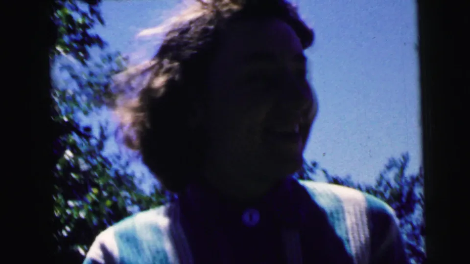 Laughing woman with curly hair and light cardigan, framed against backyard sky and trees in 8mm home movie footage, Camden, New Jersey, circa 1964.