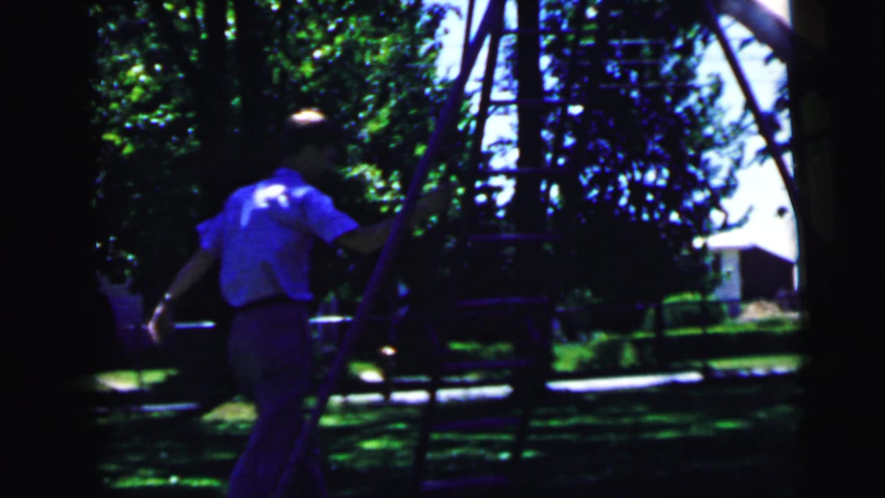 Boy in light blue shirt on a metal backyard playground structure, suburban trees and houses visible behind him, 8mm footage circa 1964, Camden, NJ.
