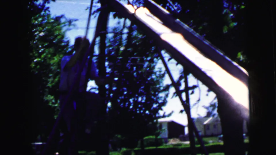 Low-angle shot along sunlit metal slide rails with strong sun flare and leafy trees, backyard setting in 8mm home movie footage circa 1964.