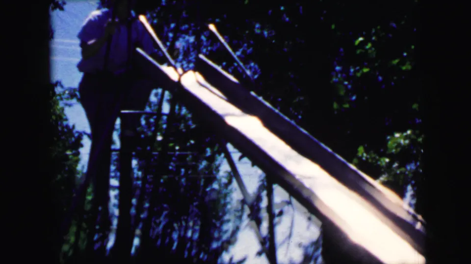 Child silhouetted at metal slide with leafy suburban trees behind, 8mm home movie footage circa 1964, Camden, New Jersey.