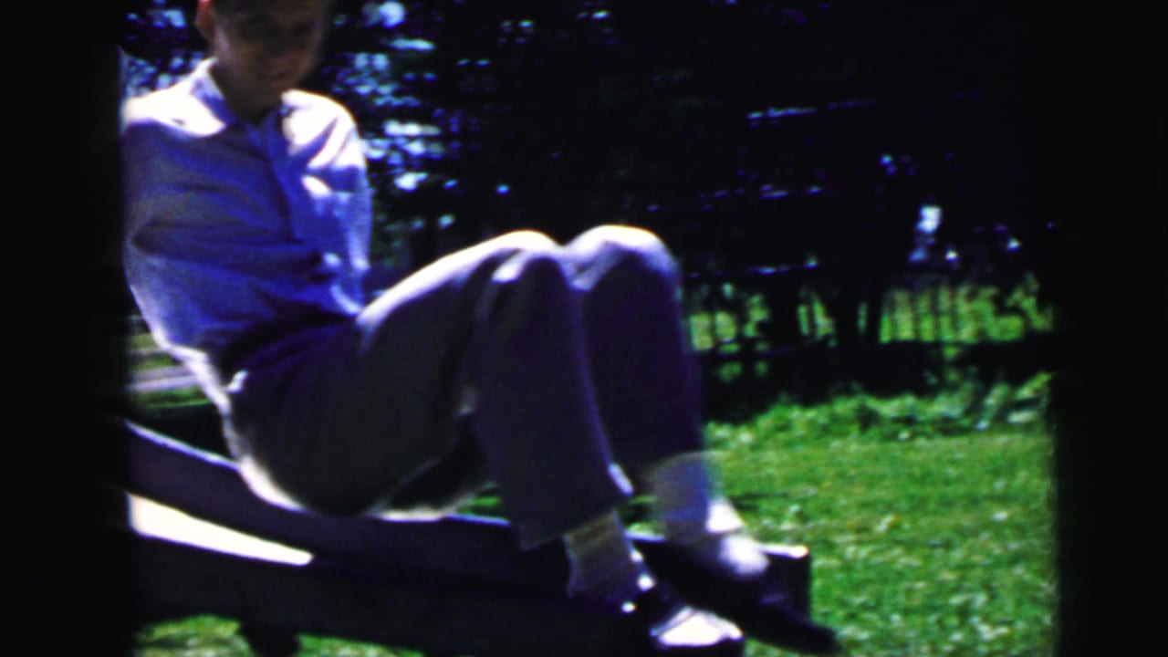 In 1964 Camden home-movie footage, a young man sits on a wooden slide beside green grass and trees.