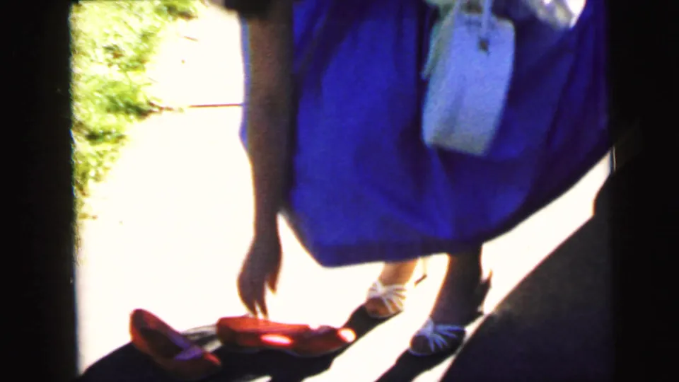 A woman in a purple dress and white shoes walks along a sunlit path, carrying a white jacket in 1964 Camden footage.