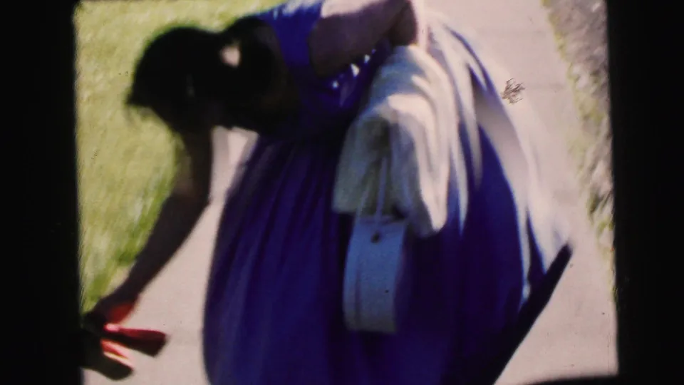 The woman in the purple dress bends near the concrete path, with grass and bright sunlight around her in the 1964 Camden clip.