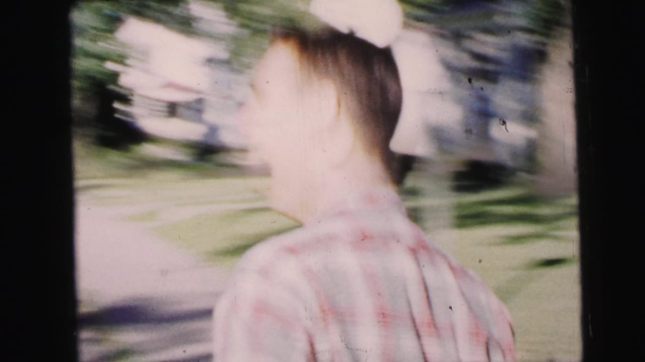 Young man in a plaid shirt walks away along a bright suburban sidewalk, with lawns and trees visible around him.