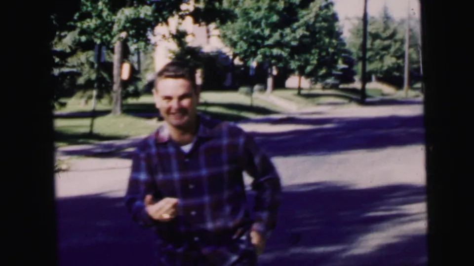 Young man in a dark plaid shirt smiles toward the camera on a sunlit Camden residential street in 1964.