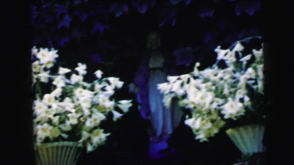 1964 8mm Camden home-movie frame showing white flower bouquets in baskets near a shadowed robed statue.