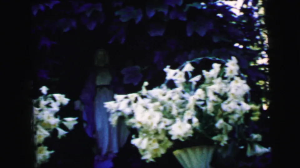 1964 home-movie frame from Camden showing white bouquets and a dimly lit robed figure in a garden.