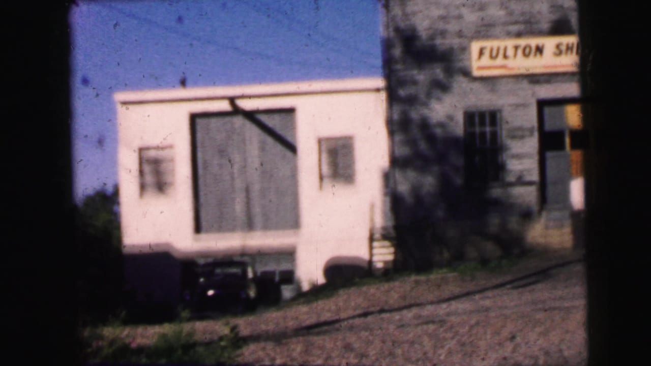 White building with a large door, parked car, sloped road, and partial Fulton sign nearby.