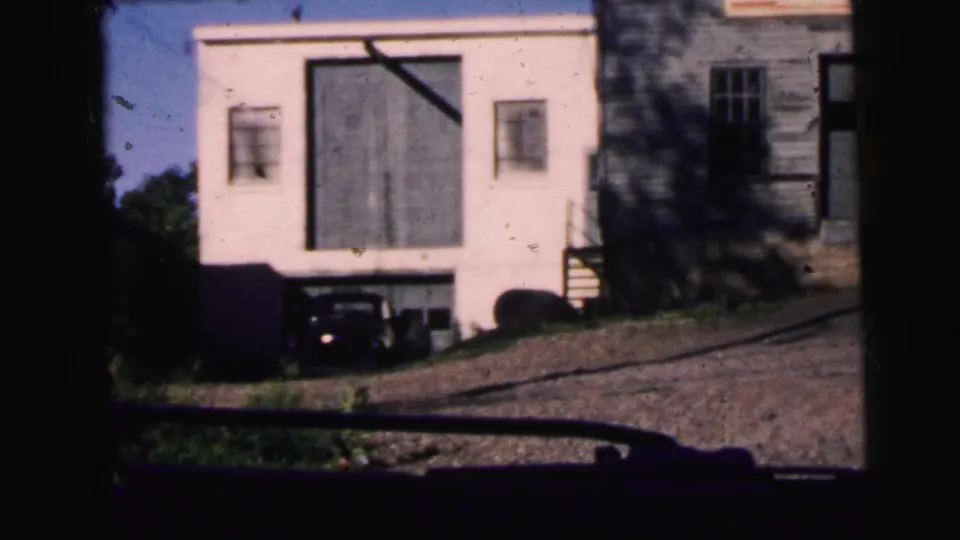 1964 Camden dashboard-view frame with a white garage-like building, older parked car, adjacent wood-sided structure, and sloped street.