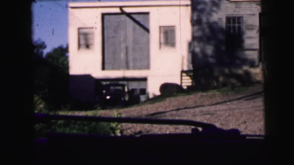 1964 Camden home-movie frame from inside a moving car, showing a white building, parked older car, neighboring wood-sided structure, and roadside slope.