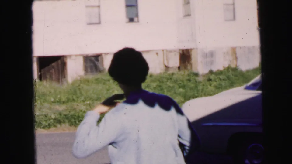 A woman in a pale cardigan is seen from behind near a parked car on a Camden residential street in 1964 footage.
