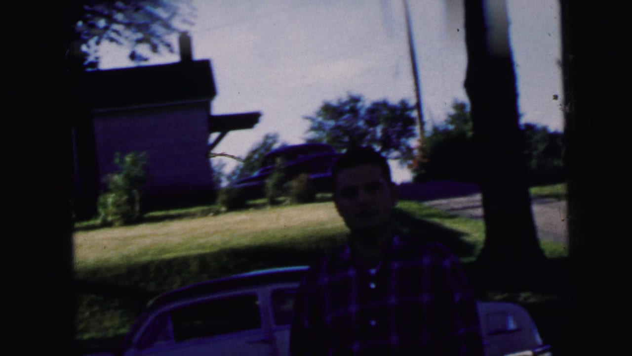 In Camden, New Jersey home-movie footage from 1964, a man in a red plaid shirt stands near a white car by trees and a house.