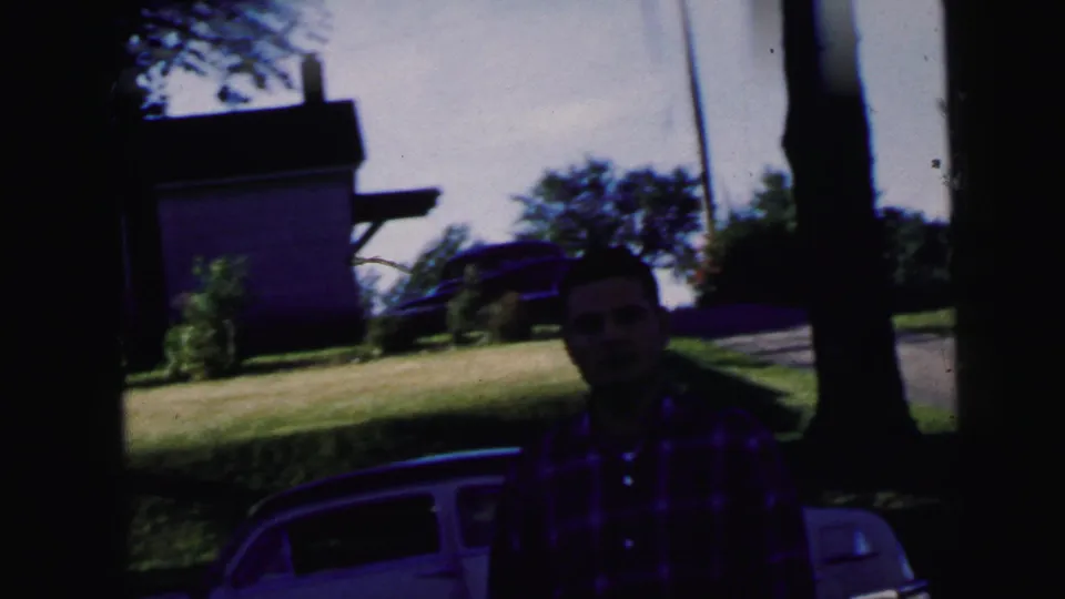 In Camden, New Jersey home-movie footage from 1964, a man in a red plaid shirt stands near a white car by trees and a house.
