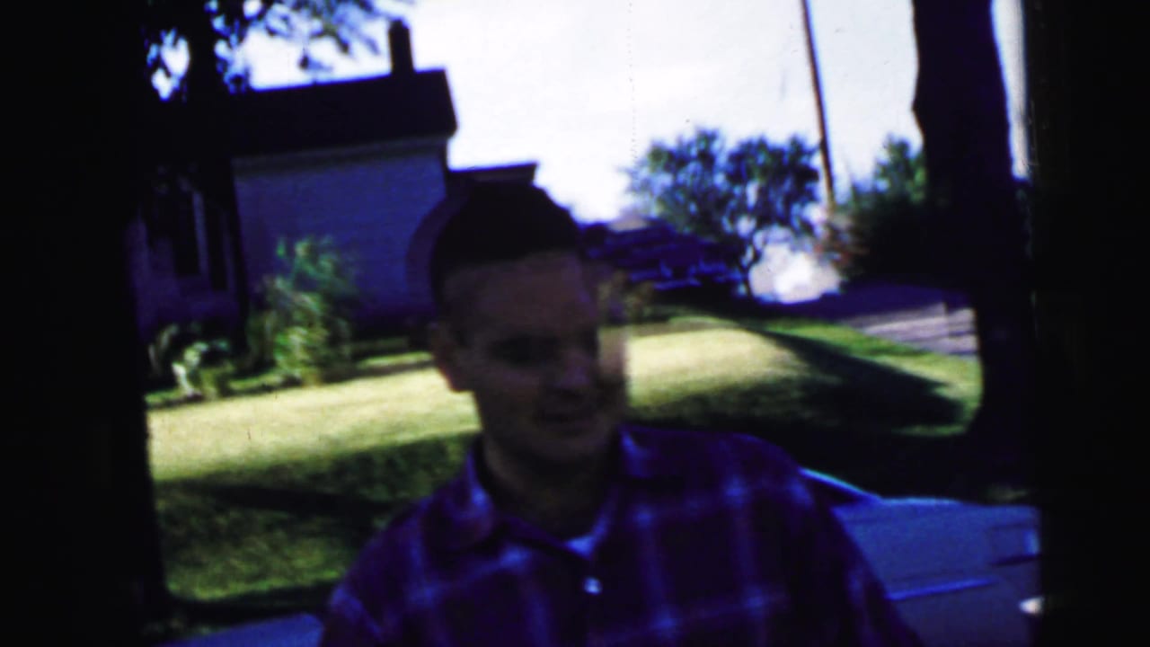The man in the red plaid shirt moves closer to the camera in a 1964 Camden residential outdoor scene.