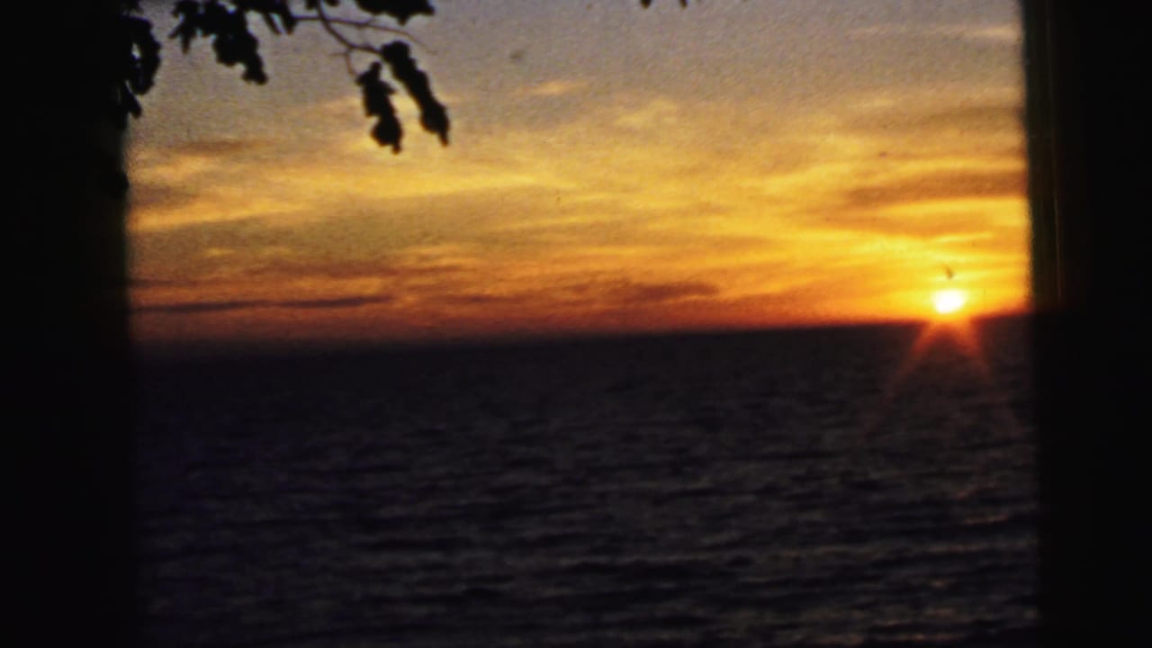 1964 8mm seaside sunset view from Camden, New Jersey, showing the sun near the horizon, rippled water, orange cloud bands, and dark foreground framing.