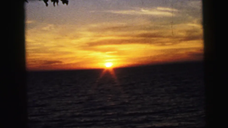 1964 8mm home-movie view of a golden sunset over the water in Camden, New Jersey, with layered clouds and a dark vertical foreground edge.