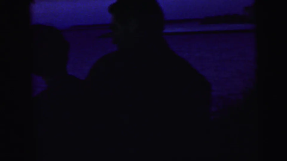 Blue-tinted home-movie frame with two adult silhouettes near the water, one larger figure centered and another at the left edge.
