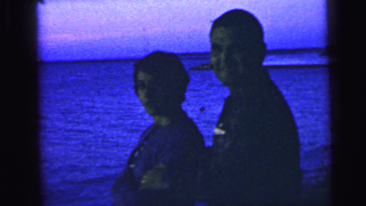 Two silhouetted adults stand close together beside water at dusk.