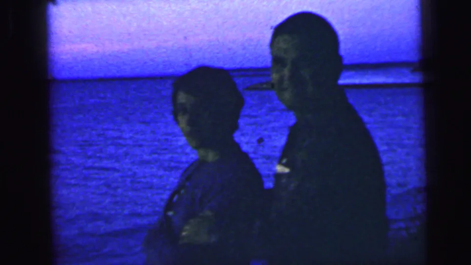 Two adults in silhouette stand together on a Camden waterfront at dusk in 1964 home-movie footage.