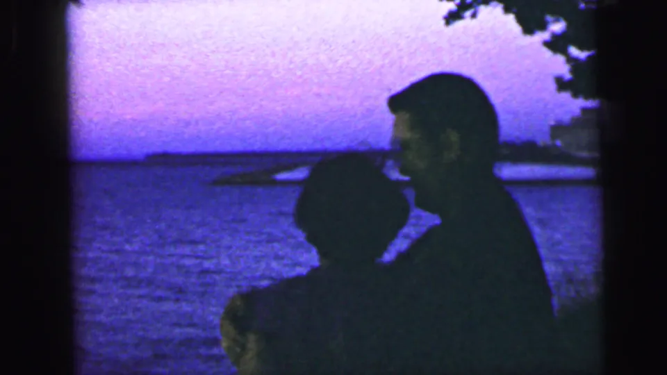 A silhouetted pair remains close on the waterfront, partly framed by dark film edges and a distant shoreline.