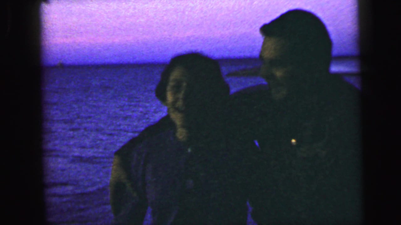 In 1964 Camden, New Jersey home-movie footage, a smiling couple stands close beside water under a purple-blue sky.