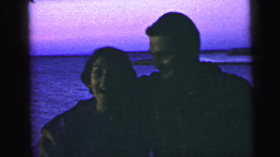 The 1964 home-movie frame shows a man embracing a smiling woman at a Camden waterfront, with blue-purple dusk light behind them.