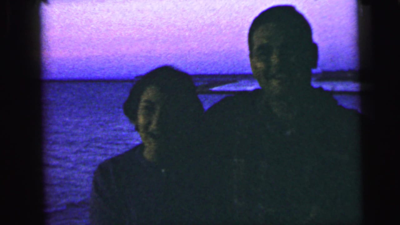 In this 1964 Camden, New Jersey home-movie frame, a couple stands close together by the water in dark silhouette.
