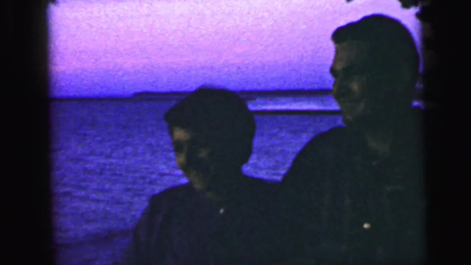 A shadowed couple faces the camera beside the water in a grainy 8mm home-movie frame from the supplied 1964 Camden setting.
