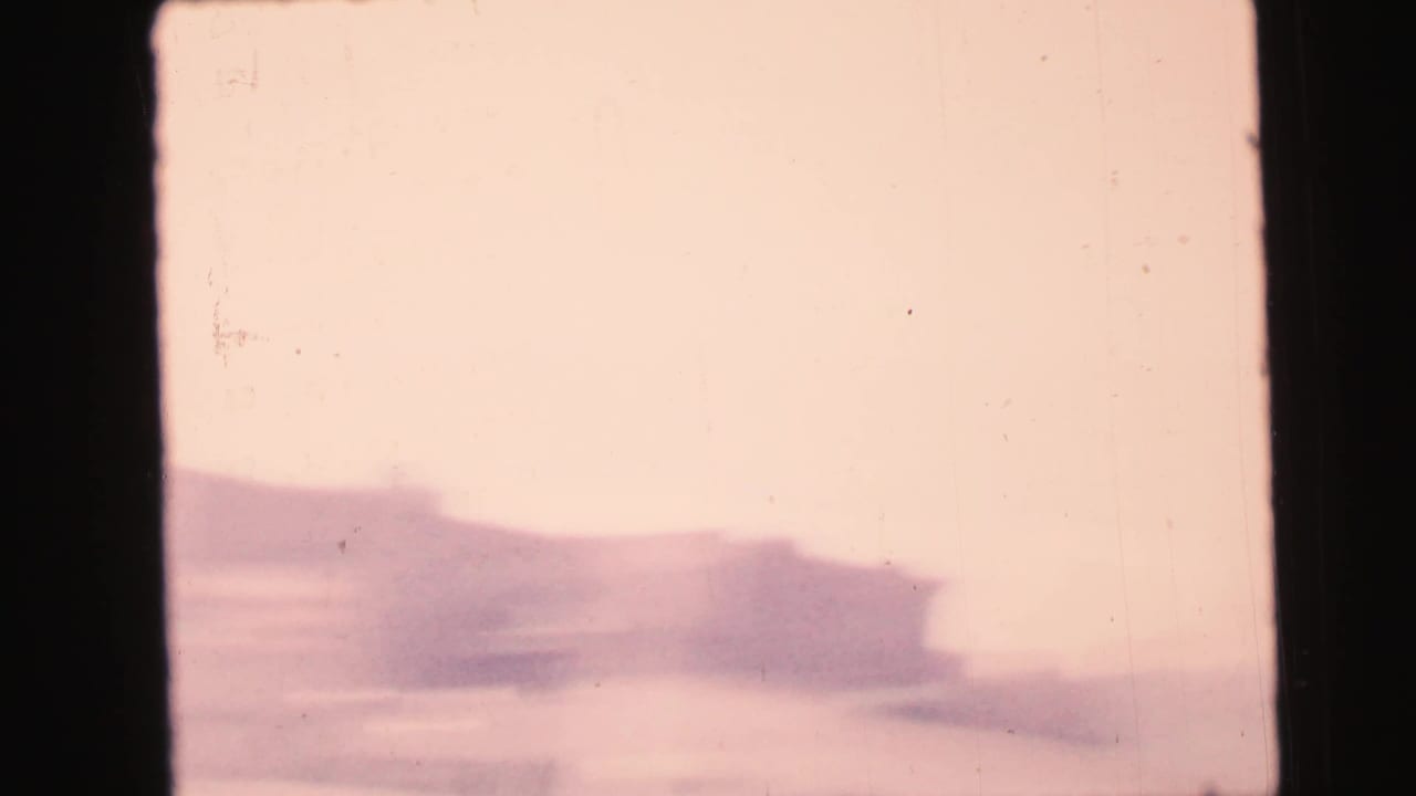 1964 Camden home-movie frame looking through dark window bars toward a soft, overexposed urban building exterior.