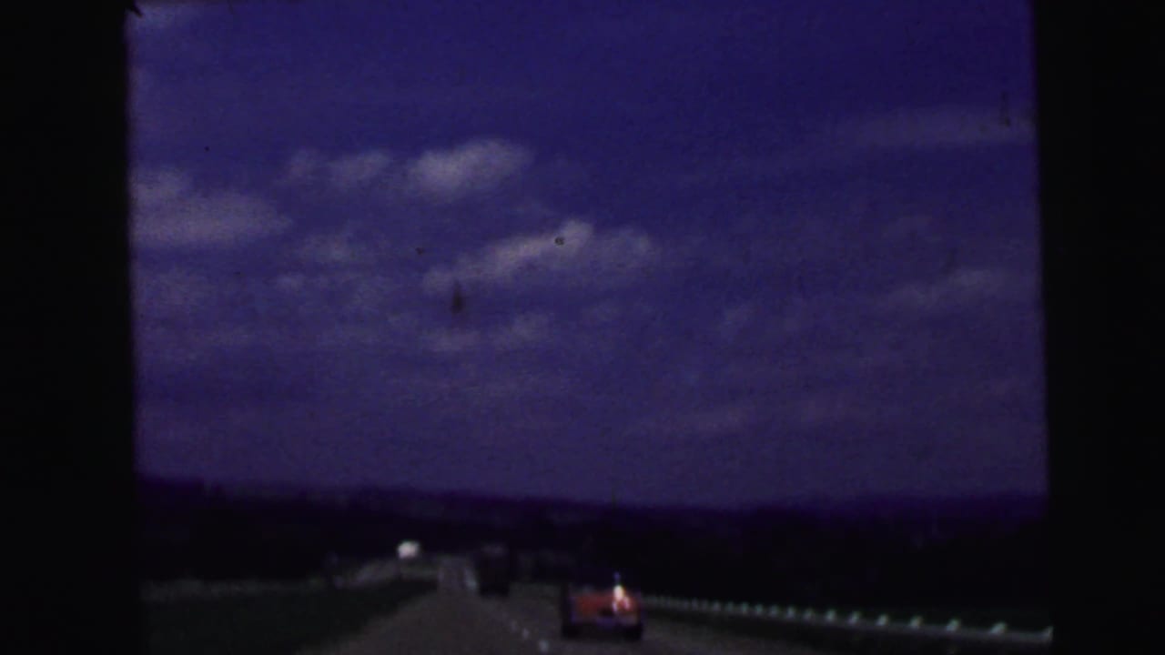 1964 home-movie road view near Camden, New Jersey, showing a highway, distant cars, guardrail, and blue sky through dark film edges.