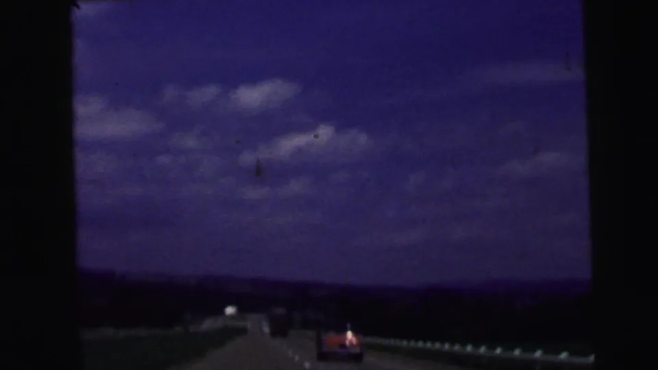 1964 home-movie road view near Camden, New Jersey, showing a highway, distant cars, guardrail, and blue sky through dark film edges.