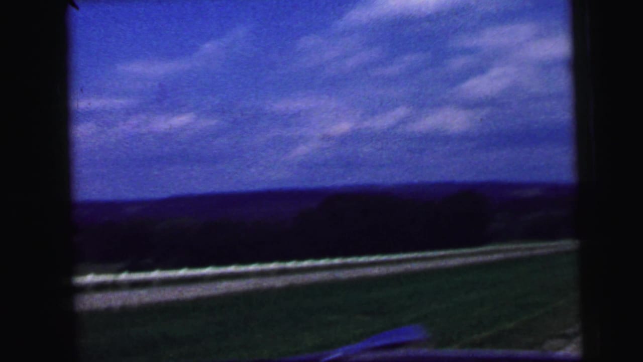 Passenger-side view from a moving vehicle, showing open grass, roadside guardrails, distant tree line, and blue sky in 1964 footage.