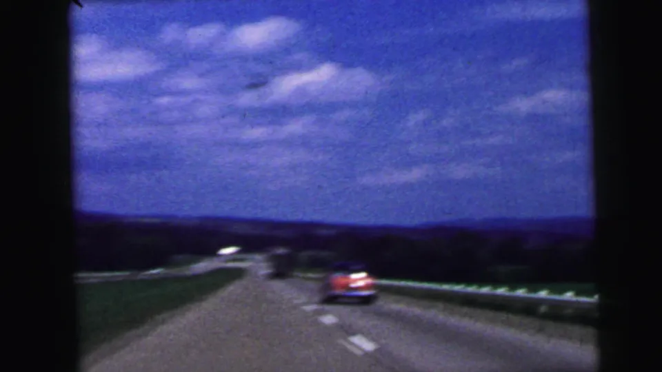 1964 8mm highway view near Camden, New Jersey, with classic cars ahead, roadside fields, guardrails, low hills, and a vivid blue sky.