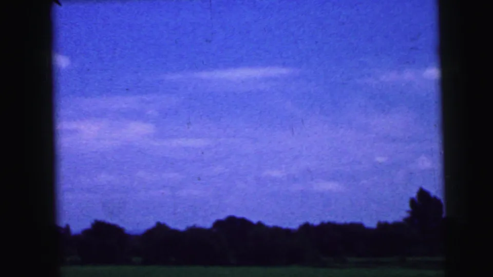 Soft 8mm landscape frame showing a bright blue sky, scattered clouds, dark tree line, and open ground, likely filmed during the road trip.