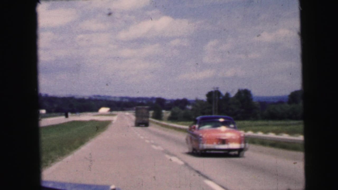 The road scene shows a pink midcentury car, a truck, grassy shoulders, and cloudy sky in Camden-area 1964 footage.