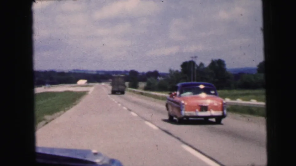 A passenger-shot 8mm frame shows the rear of a pink sedan beside a truck on a divided New Jersey highway in 1964.