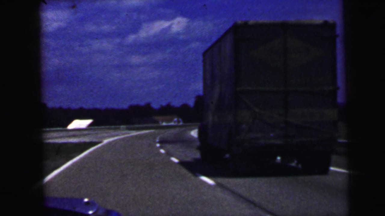 1964 8mm home-movie frame from the Camden, New Jersey area showing a large truck ahead on a curving highway under a blue sky, with the car interior partly visible.