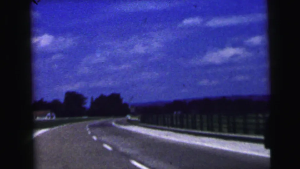 1964 8mm frame from a moving vehicle near Camden, New Jersey, showing an open curving highway, dashed lane markings, roadside barrier, trees, and a saturated blue sky.