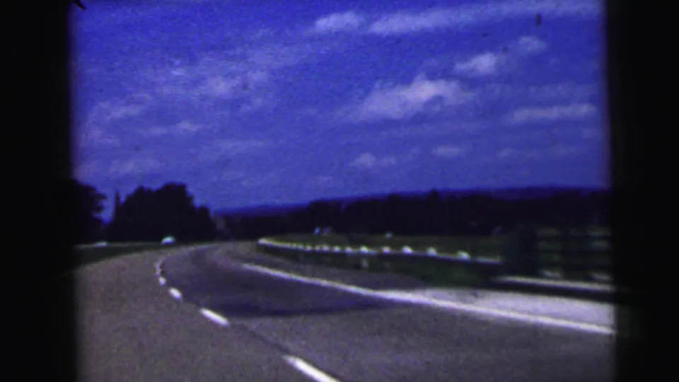 1964 8mm home-movie view in the Camden, New Jersey area showing a curving highway bordered by guardrails, fields or low vegetation, distant trees, and scattered clouds.