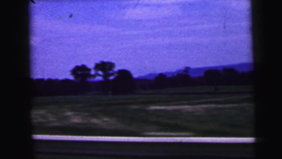 1964 home-movie view near Camden, New Jersey, showing fields, silhouetted trees, and a roadside edge from a moving car.