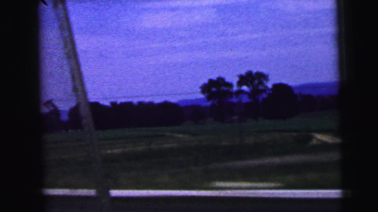 Dark train window frame looking onto green land, trees, and distant hills under a blue-purple sky.