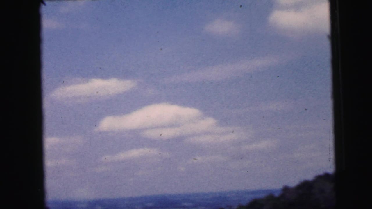 Blue sky and soft white clouds over a distant horizon, framed by the moving vehicle or film edge in 1964 Camden footage.