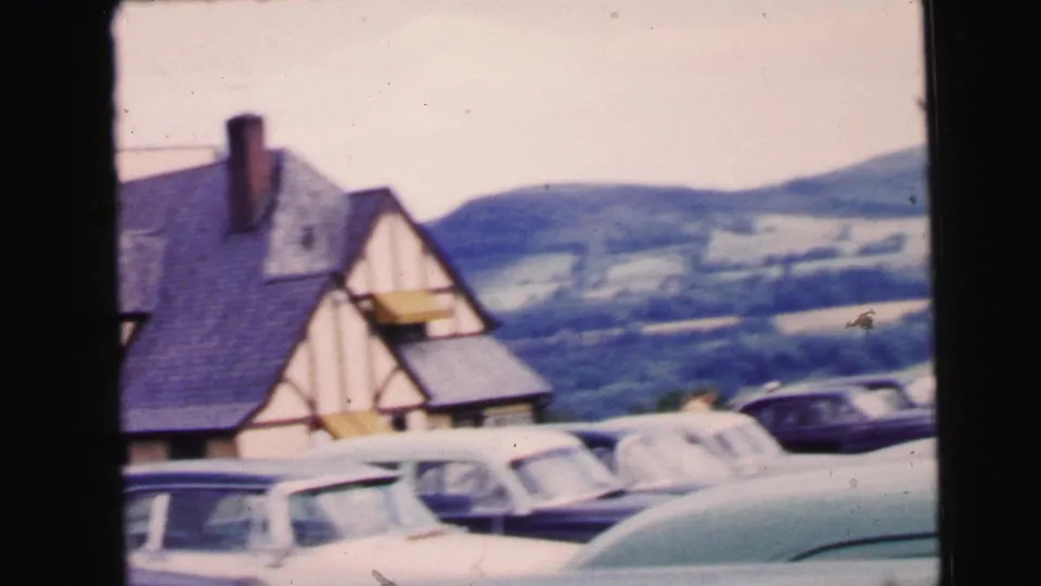 Classic cars sit near a Tudor-style roadside building with hills beyond in the 1964 home-movie clip.