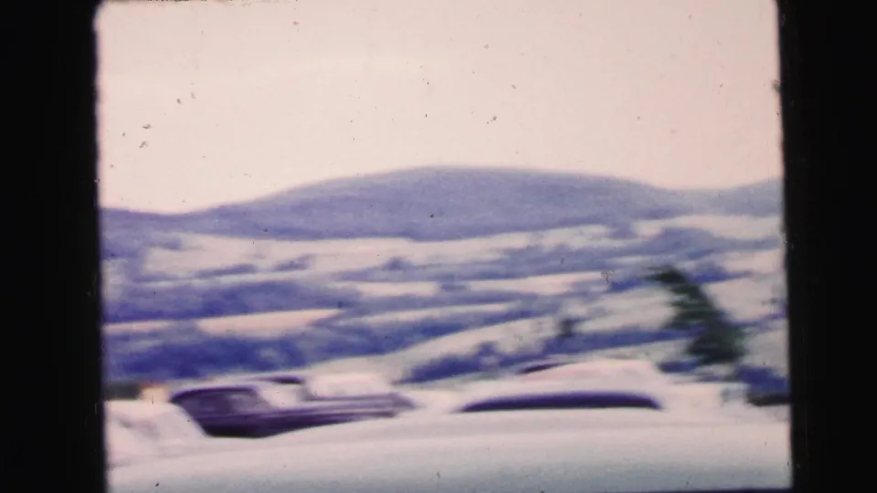 A soft 8mm view shows parked mid-century cars with rolling hills in the background, within the 1964 travel footage.