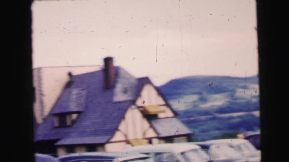 1964 Camden, New Jersey 8mm frame showing a steep gabled roof, chimney, parked cars, and low hills beyond.