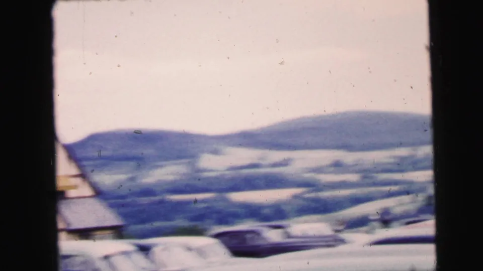 1964 Camden, New Jersey frame showing snow-covered ground, parked automobiles, and soft low hills beyond a partial roofline.