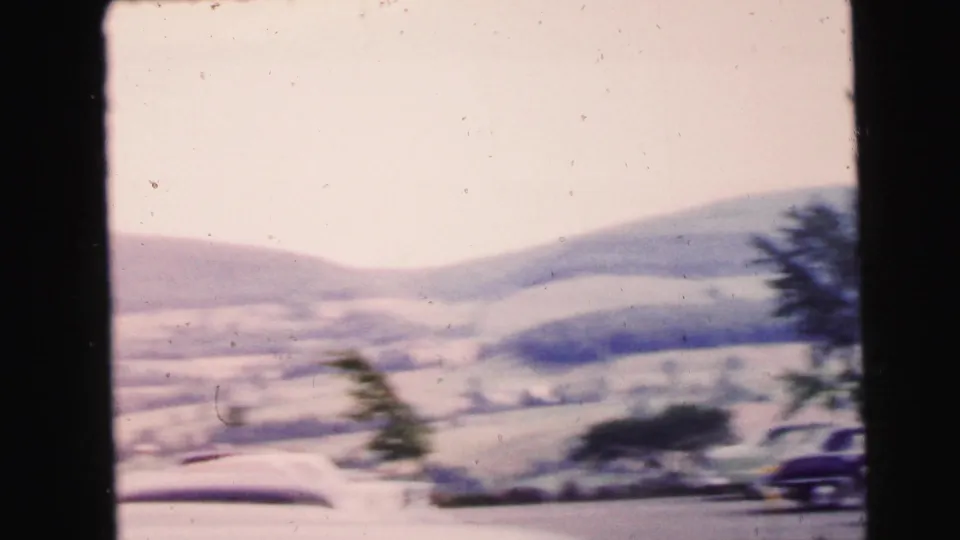 1964 home-movie view of a car-show parking area near Camden, New Jersey, seen through dark frame edges with hills beyond.