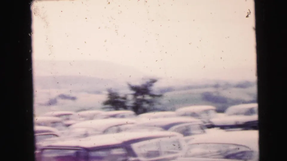 Soft 8mm frame of a Camden, New Jersey car-show parking area with blurred cars, a tree, and pale hills in the distance.